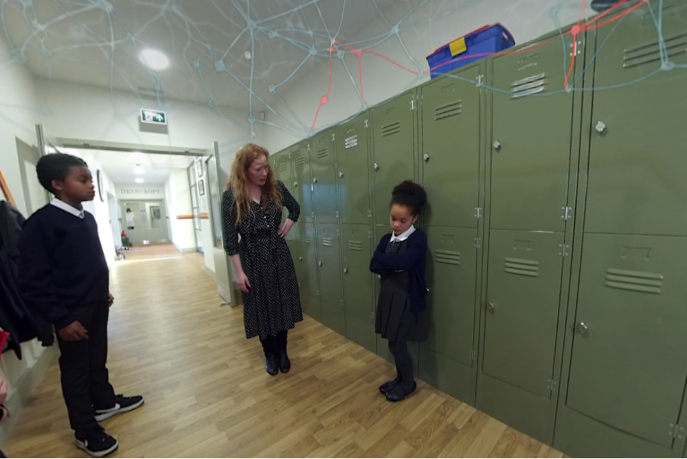 A still taken from the Cornerstone VR vulnerabilities in adolescence VR films. It shows a young girl, teacher and young boy, in a school corridor by lockers.